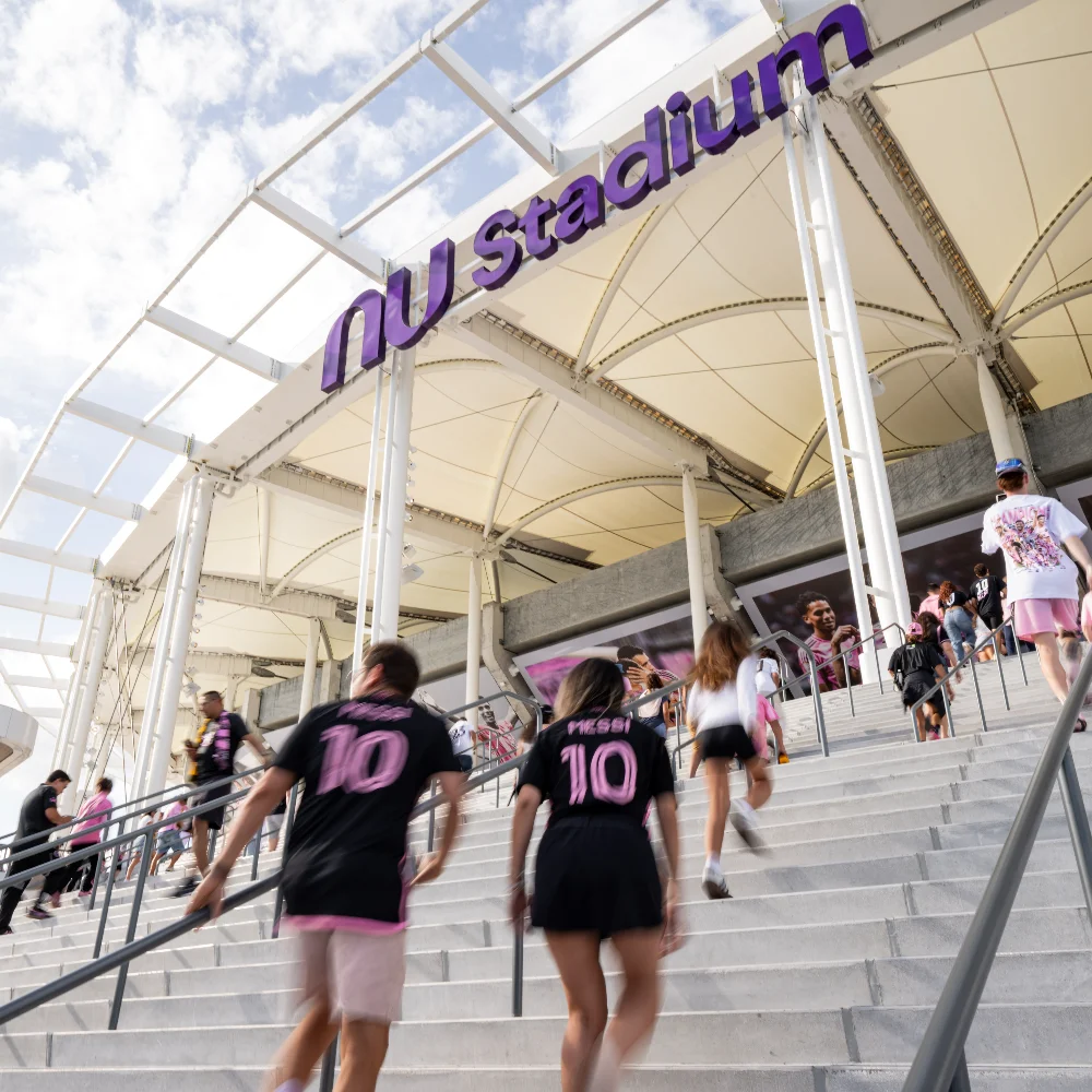 Visitors wearing Inter Miami jerseys walk up the entrance stairs to NU Stadium under a white canopy structure.
