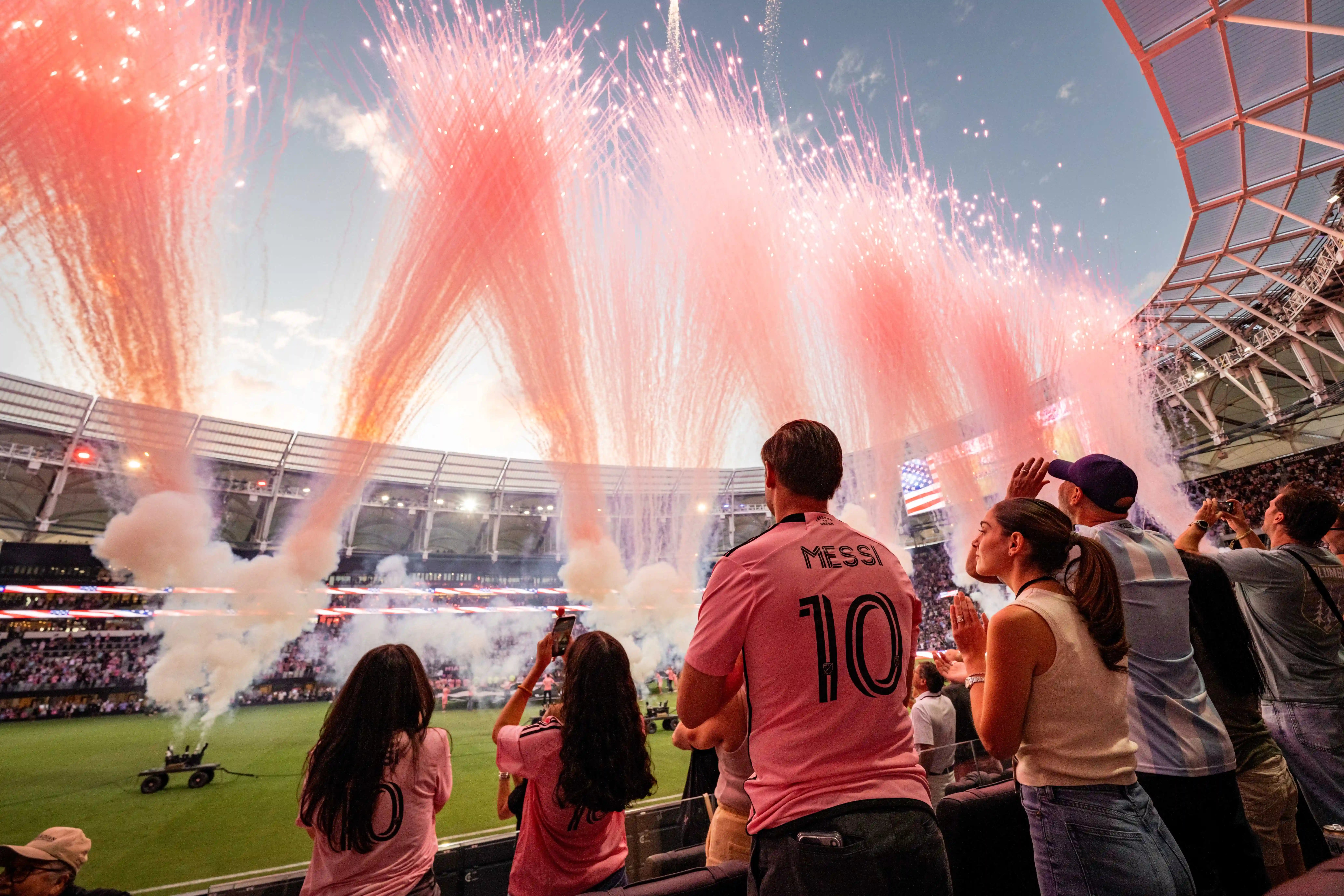 Fans wearing Inter Miami jerseys watch fireworks and pre-match celebrations from the stands inside a soccer stadium.
