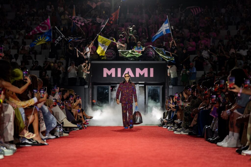 Model walks down a red runway at an Inter Miami CF fashion show as fans wave flags and take photos.